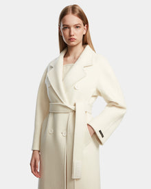 A woman with straight, light brown hair wears the Rudsak KOURT W - 8124702 Iceland, a long white women’s belted wool coat with wide lapels, made from a luxurious wool and cashmere blend, standing against a plain light gray background.