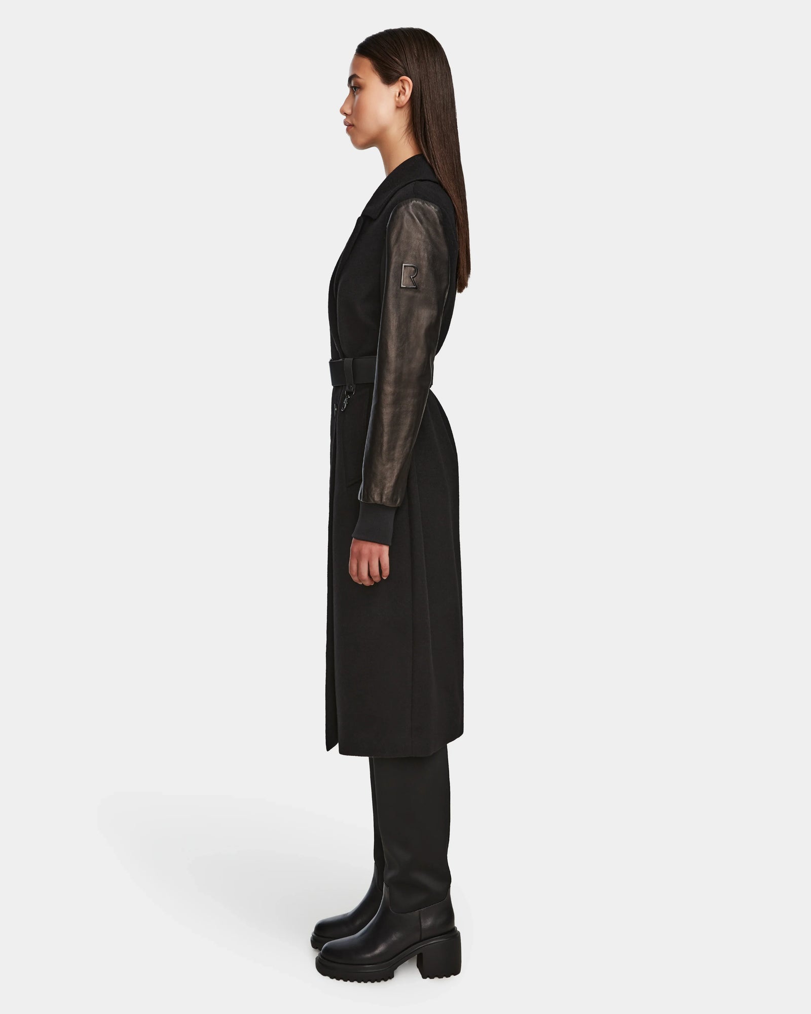 A woman in profile wears the MARYLIN - 8124520 Black womens wool coat by Rudsak, featuring leather sleeves and a belted waist, paired with knee-high black boots against a plain white background.