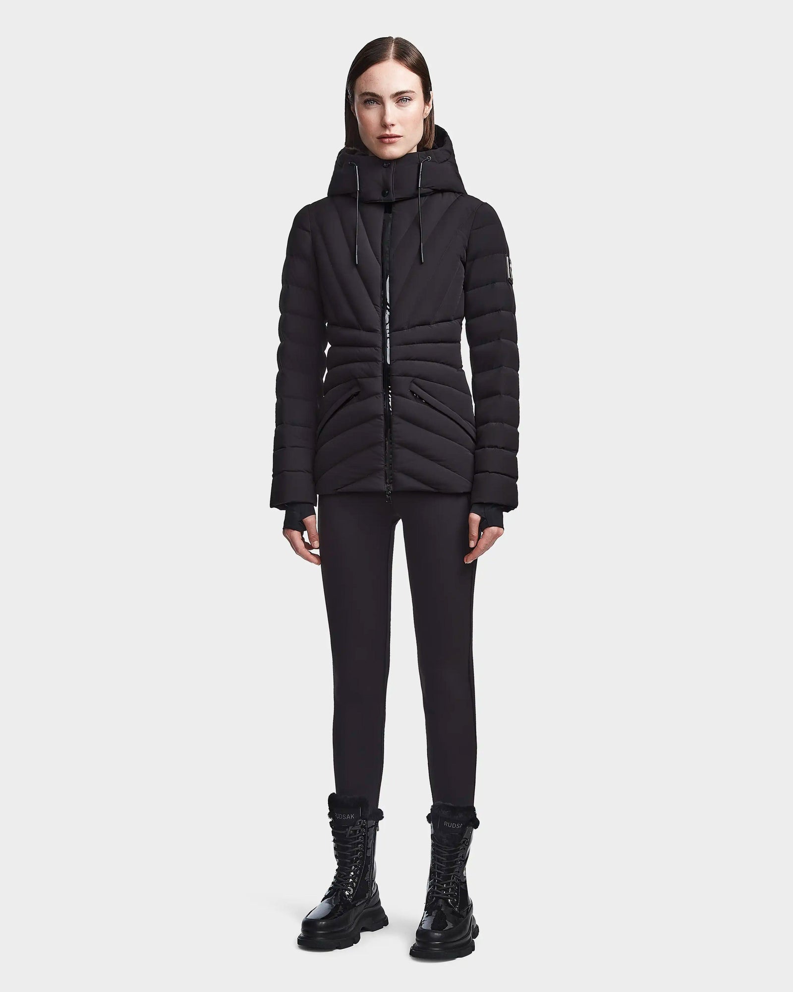 A woman faces forward against a light gray background, wearing the Rudsak HANNA - 8123562 Black winter down jacket with RDS certified down, paired with black leggings and black lace-up boots.