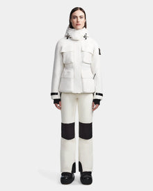 A woman stands facing forward in the Rudsak LUX - 8123618 Iceland ski jacket with large front pockets and a hood, paired with white pants with black knee patches and black boots, set against a plain light gray background.