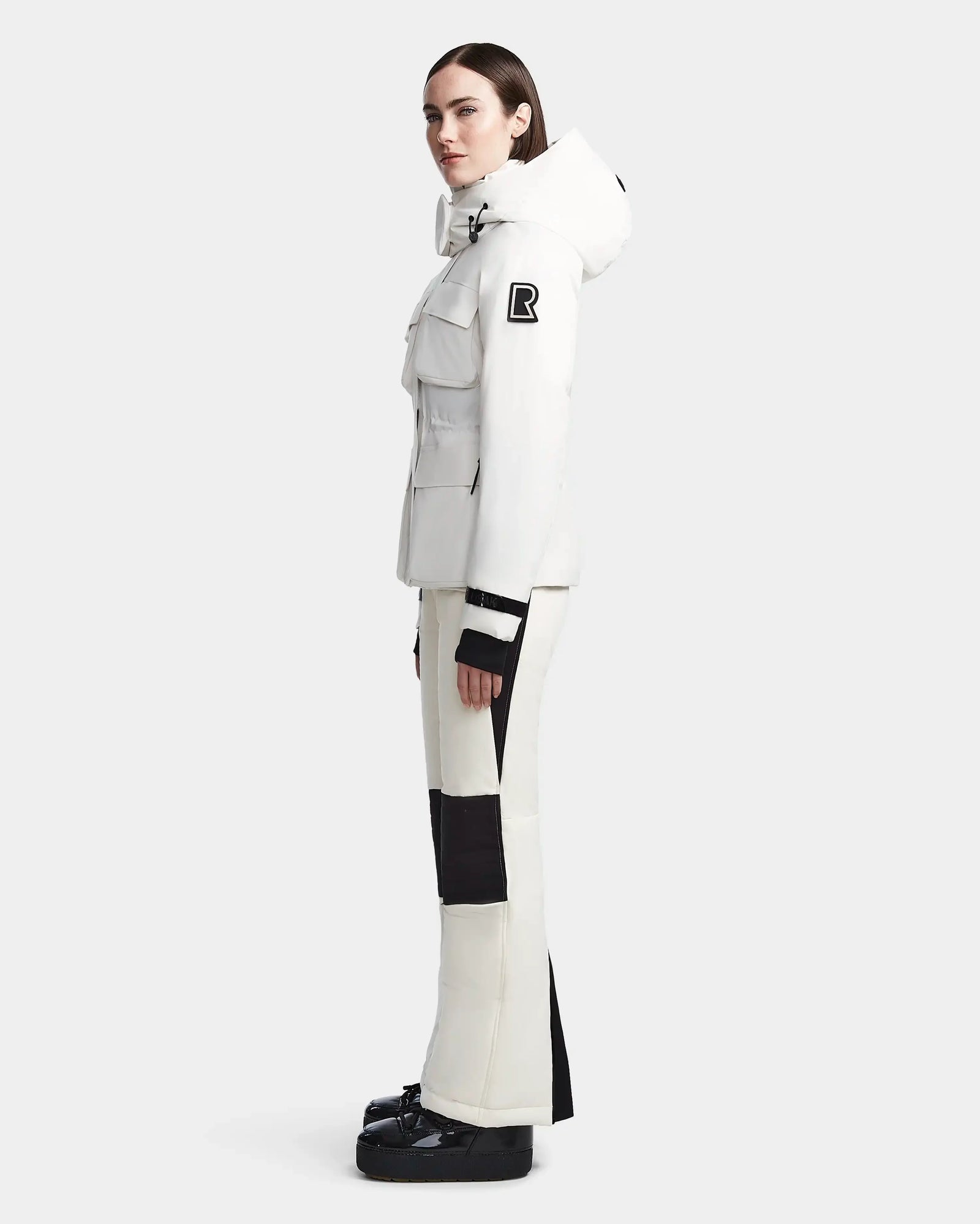 A woman stands in profile wearing the Rudsak LUX - 8123618 Iceland white hooded women’s ski jacket with black accents and flap pockets, paired with matching pants and black boots. The waterproof jacket combines stylish design with protective performance.