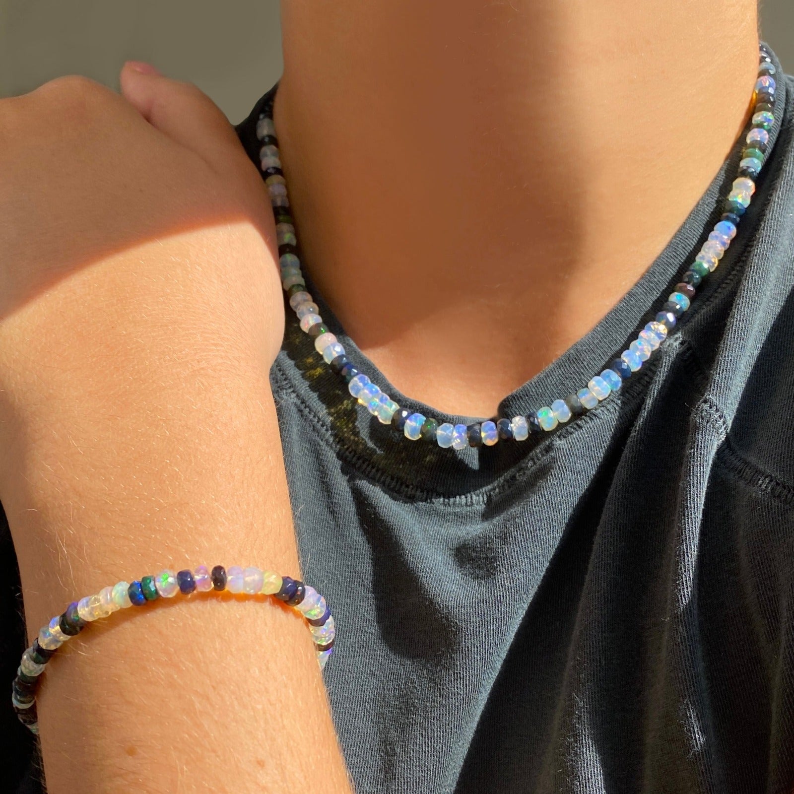 Shimmering beaded necklace made of faceted opals in shades of fiery pastels, grey, teal, and black on a gold linking ovals clasp. Layered on a neck with matching bracelet on wrist