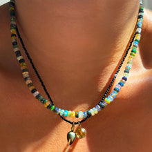Shimmering beaded necklace made of faceted opals in shades of green, blue, purple, and white on a gold linking ovals clasp styled on a neck layered with black spinel slim gemstone and charms