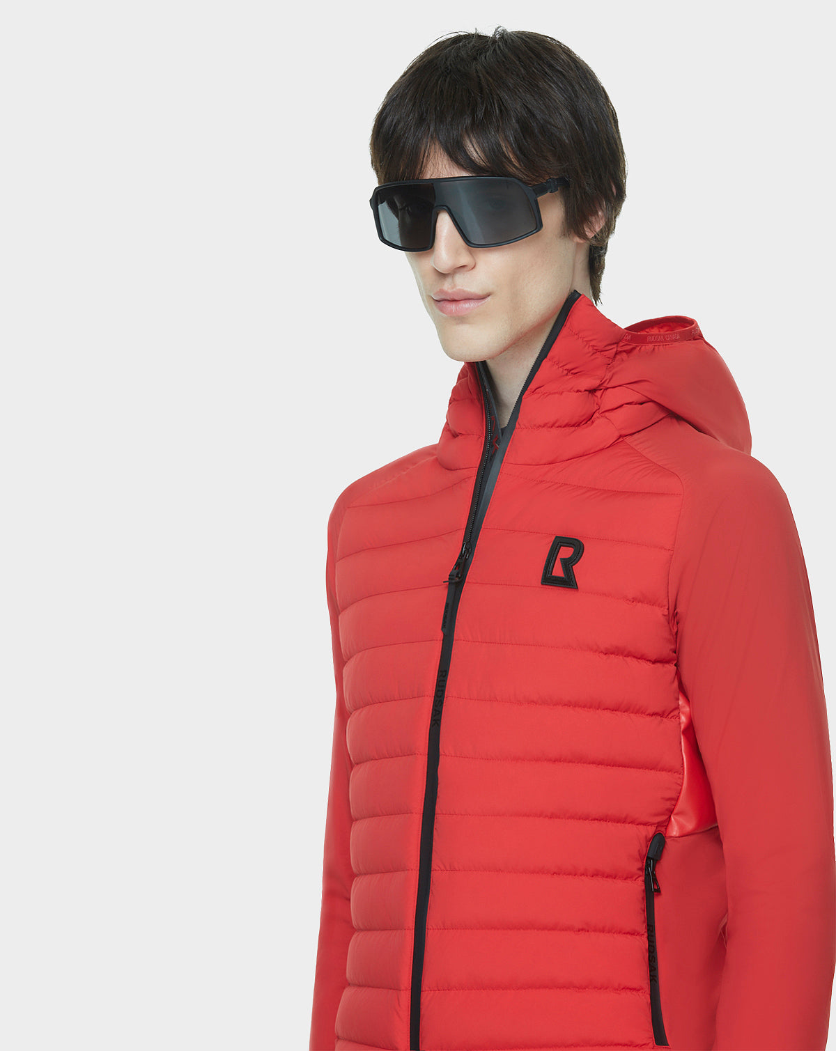 A young person models the RUDSAK JARRY - 6122007 POPPY RED water-resistant jacket with a black zipper and logo, paired with large black sunglasses, standing against a plain white background.
