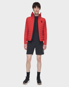 A young man stands facing forward against a plain white background, wearing the RUDSAK JARRY - 6122007 POPPY RED windbreaker over a black shirt, black shorts, black socks, and black sneakers.