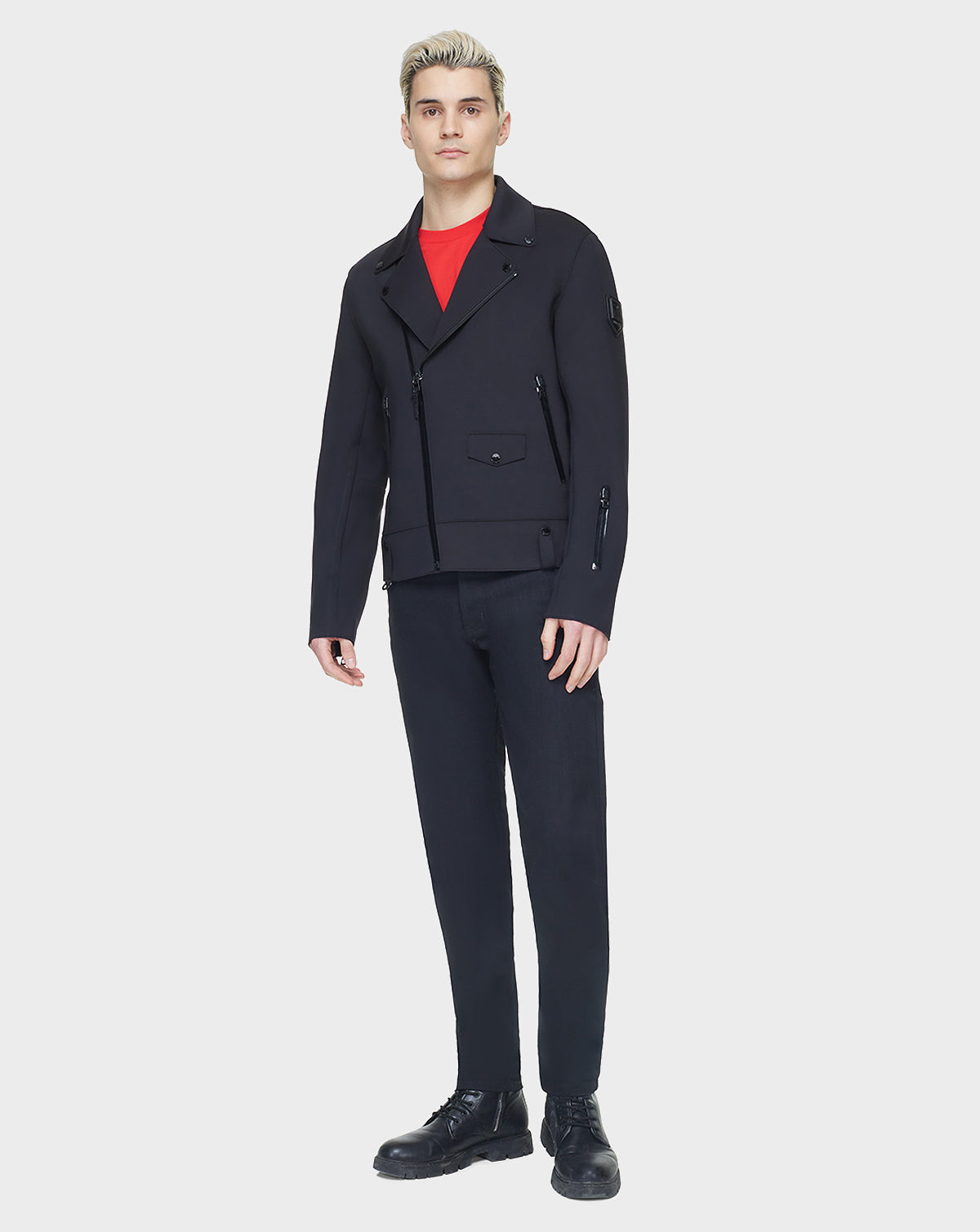 A person with short blonde hair wears the RUDSAK JUNE - 6122005 BLACK biker jacket with black pants, black shoes, and a bright red shirt, standing against a plain white background.