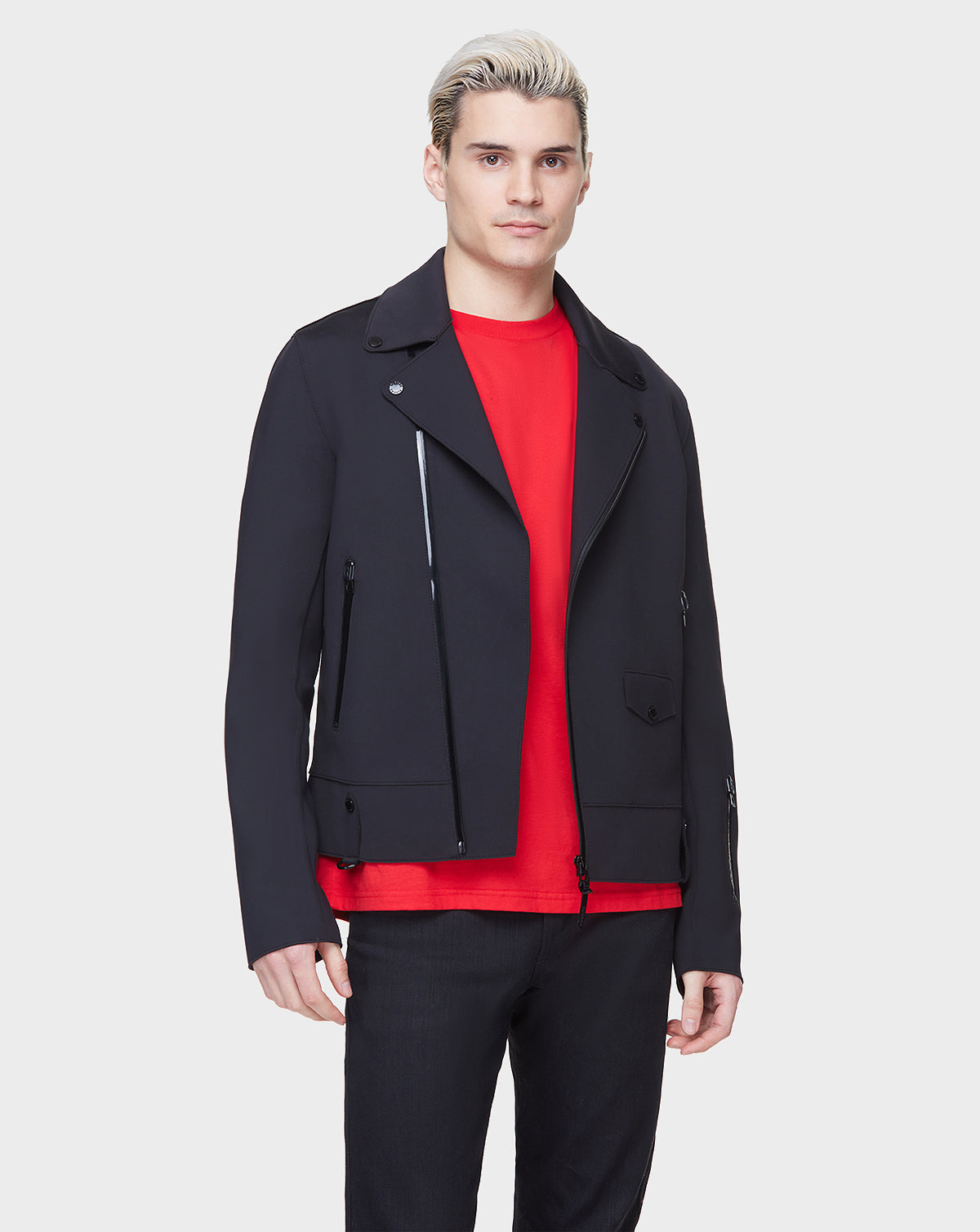 A man with short, light blond hair models the RUDSAK JUNE - 6122005 BLACK biker jacket over a bright red shirt and black pants against a plain light gray background.