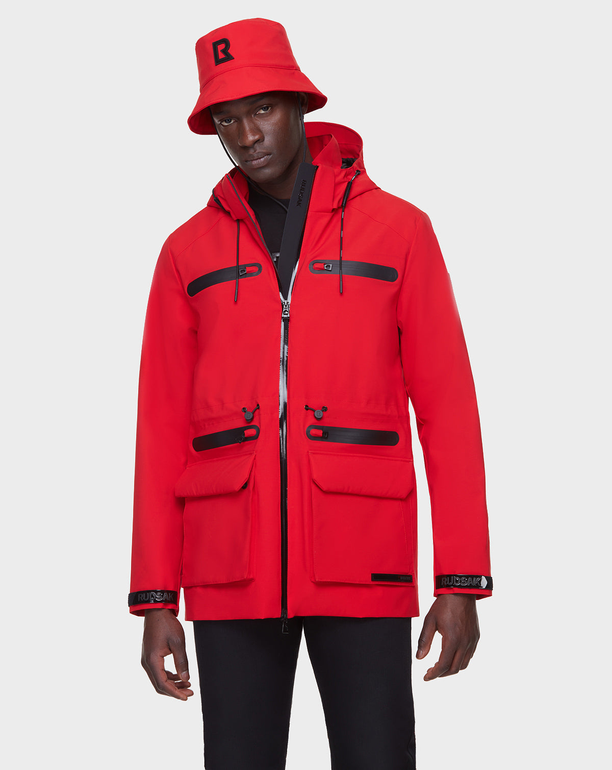 A man wears the RUDSAK JACY - 6122000 trench coat in poppy red with black zippers and front pockets, paired with a matching red bucket hat with a black R logo, standing against a plain light background.