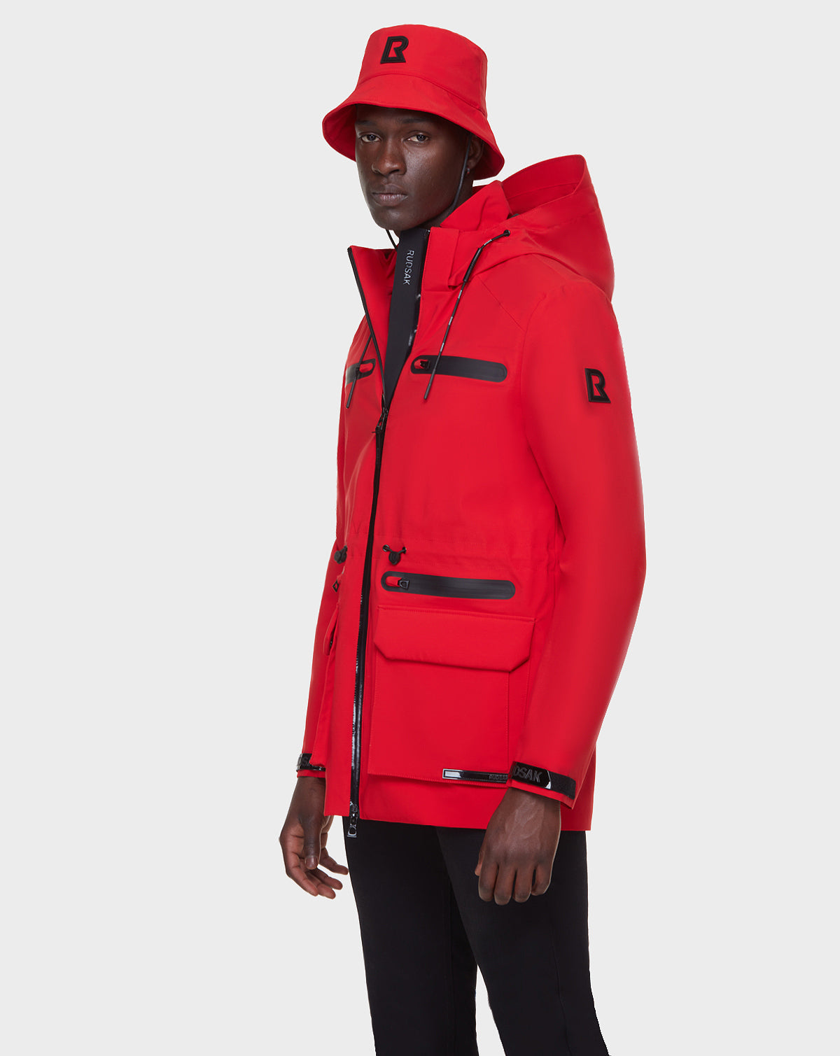 A man wears the RUDSAK JACY - 6122000 POPPY RED mens rainwear trench with black zippers and a matching red bucket hat, standing against a plain light background.