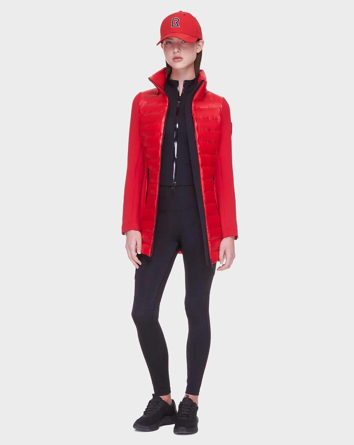 A woman faces forward in a Rudsak MERINA - 8121006 POPPY RED quilted down jacket, red baseball cap, black zip-up top, leggings, and sneakers against a plain light background.