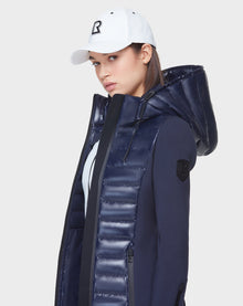 A woman in the Rudsak MARRY - CA EN L121003 DEEP BLUE puffer jacket with a hood and zippered pockets and a white baseball cap, looking to the side against a plain light background.