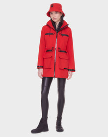 A person stands against a plain background wearing the Rudsak CAMY - 8122002 POPPY RED water-resistant coat with multiple pockets and removable hood, paired with a red bucket hat, black leggings, and black boots.