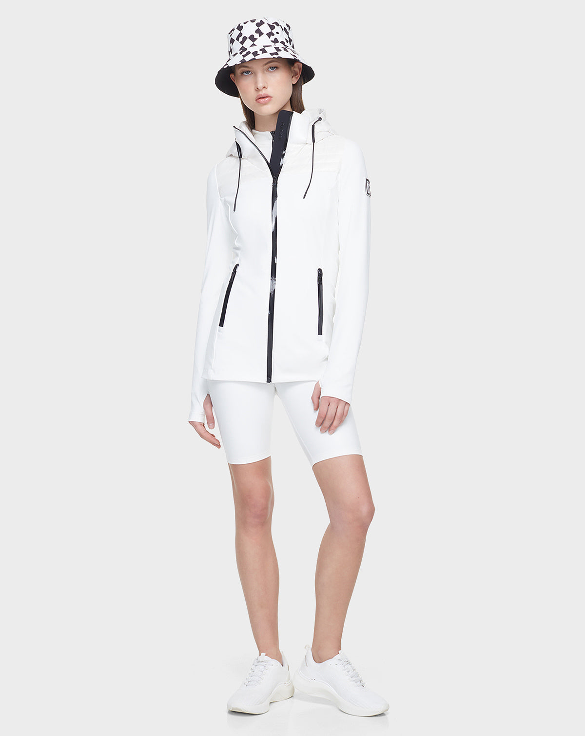 A person wears the Rudsak CAMIRA - 8122011 WHITE womens activewear jacket, paired with white cycling shorts, sneakers, and a patterned bucket hat against a light gray background—perfect for stylish outdoor workouts.